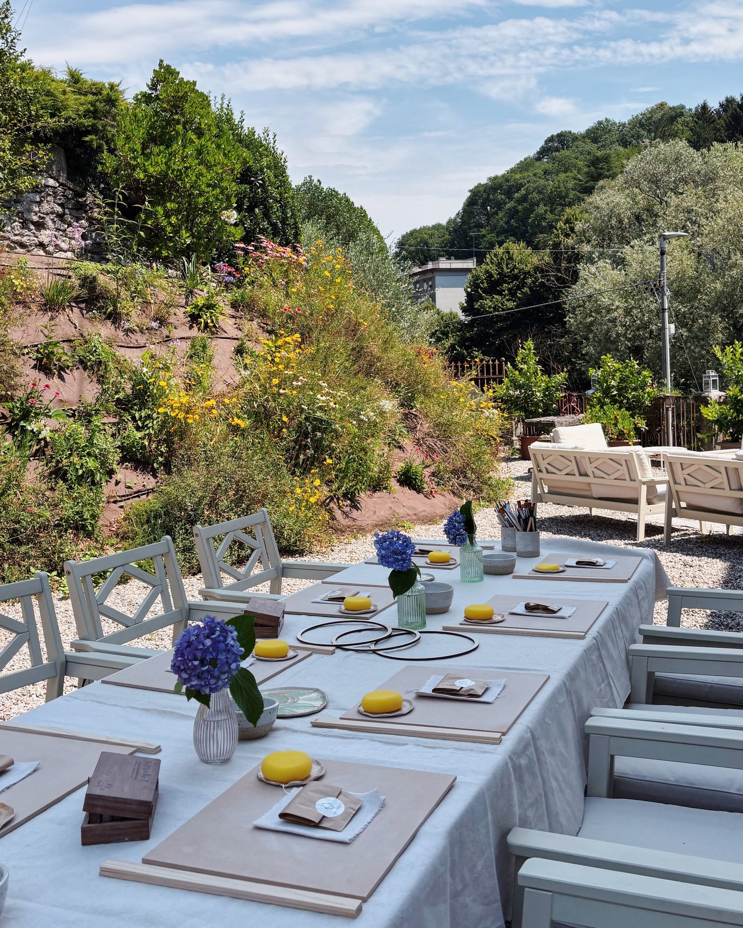 An outdoor dining setup with a long table covered in a white tablecloth, set with place settings, small yellow lemons, and purple flower arrangements, surrounded by white chairs. The scene is set in a garden with a stone wall, greenery, and a blue sky with clouds in the background.