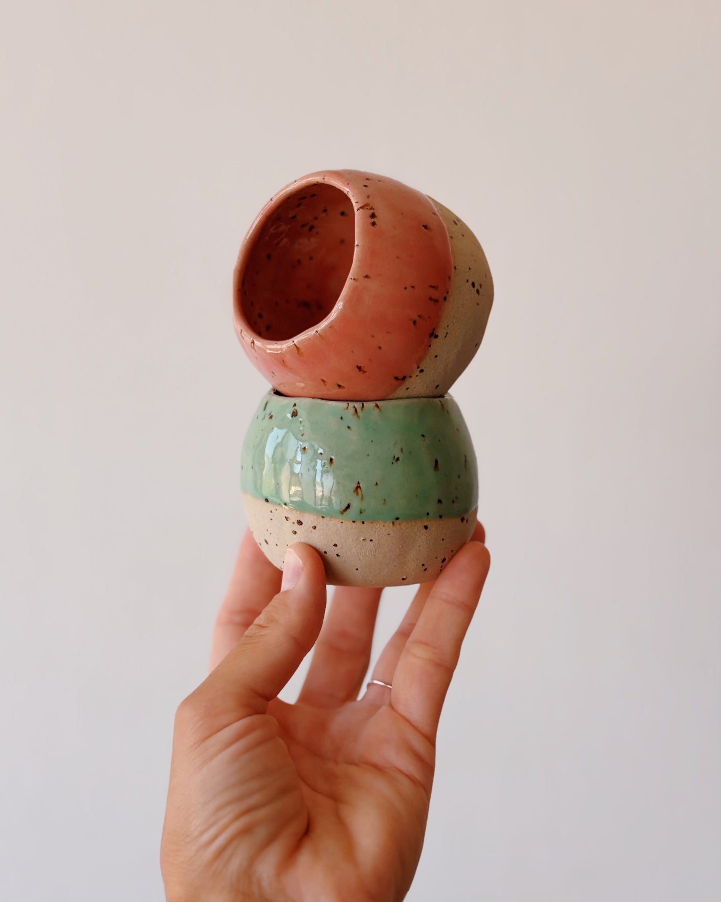 A hand holding two small handmade ceramic coffee cups with a stacked, rounded design in terracotta, sage green, and cream with black speckles, against a plain light gray background.