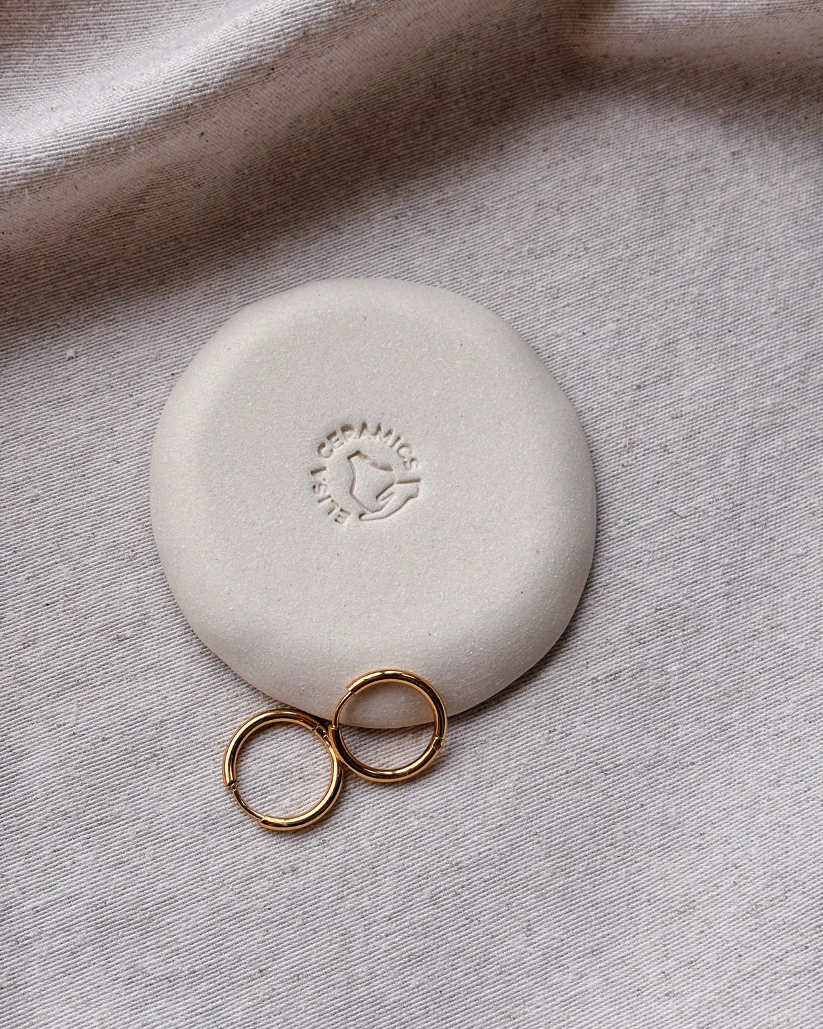 A round, matte off-white ceramic or stone jewelry box lid with an embossed logo, shown on a textured light gray fabric next to two small gold rings.