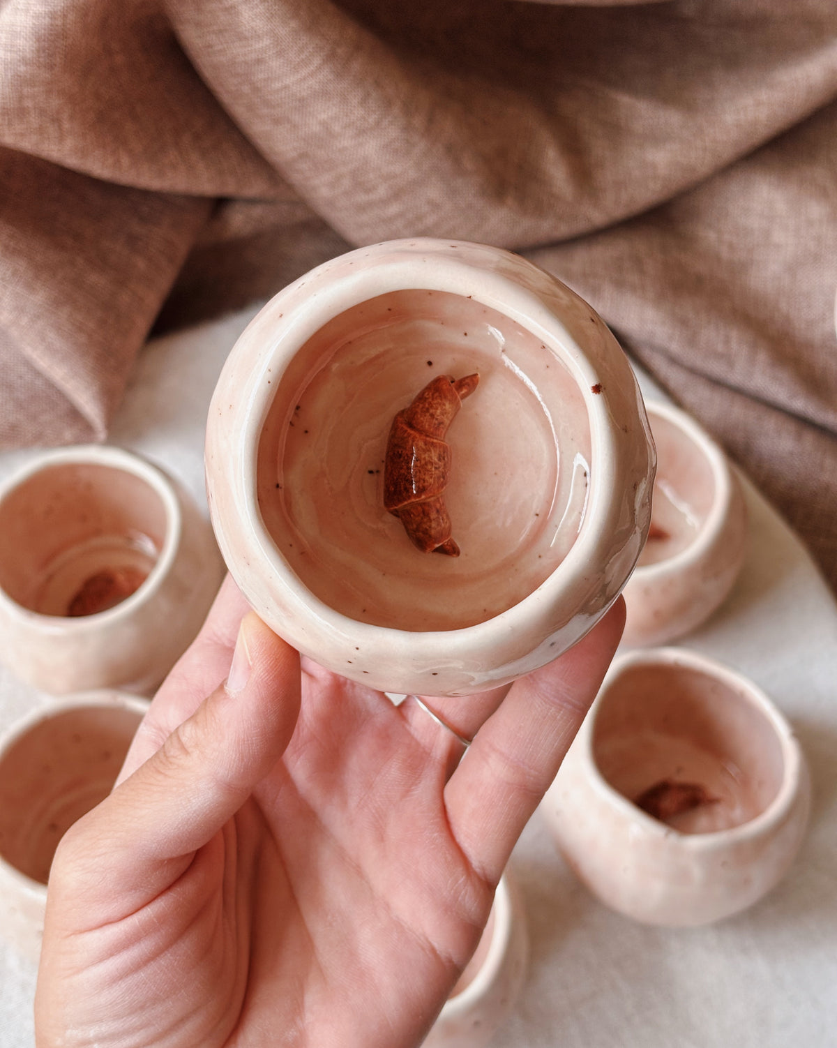 A hand holds a small handmade ceramic cup with a glossy light pink-beige glaze, showing a small croissant at the bottom. Several matching cups are arranged in the background on a light surface with a draped brown fabric backdrop.