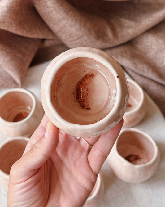 A hand holds a small handmade ceramic cup with a glossy light pink-beige glaze, showing a small croissant at the bottom. Several matching cups are arranged in the background on a light surface with a draped brown fabric backdrop.