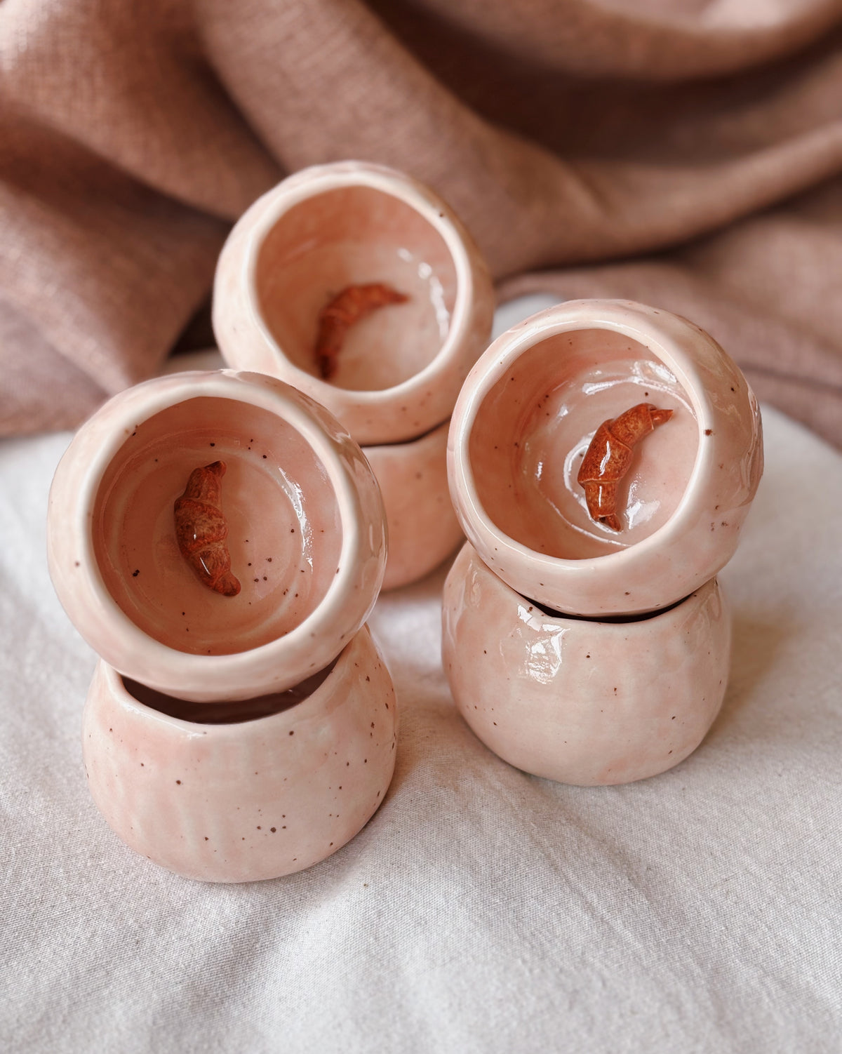 Four small stacked ceramic cups in a dusty pink speckled glaze, arranged on a light fabric surface with a soft brown cloth in the background.