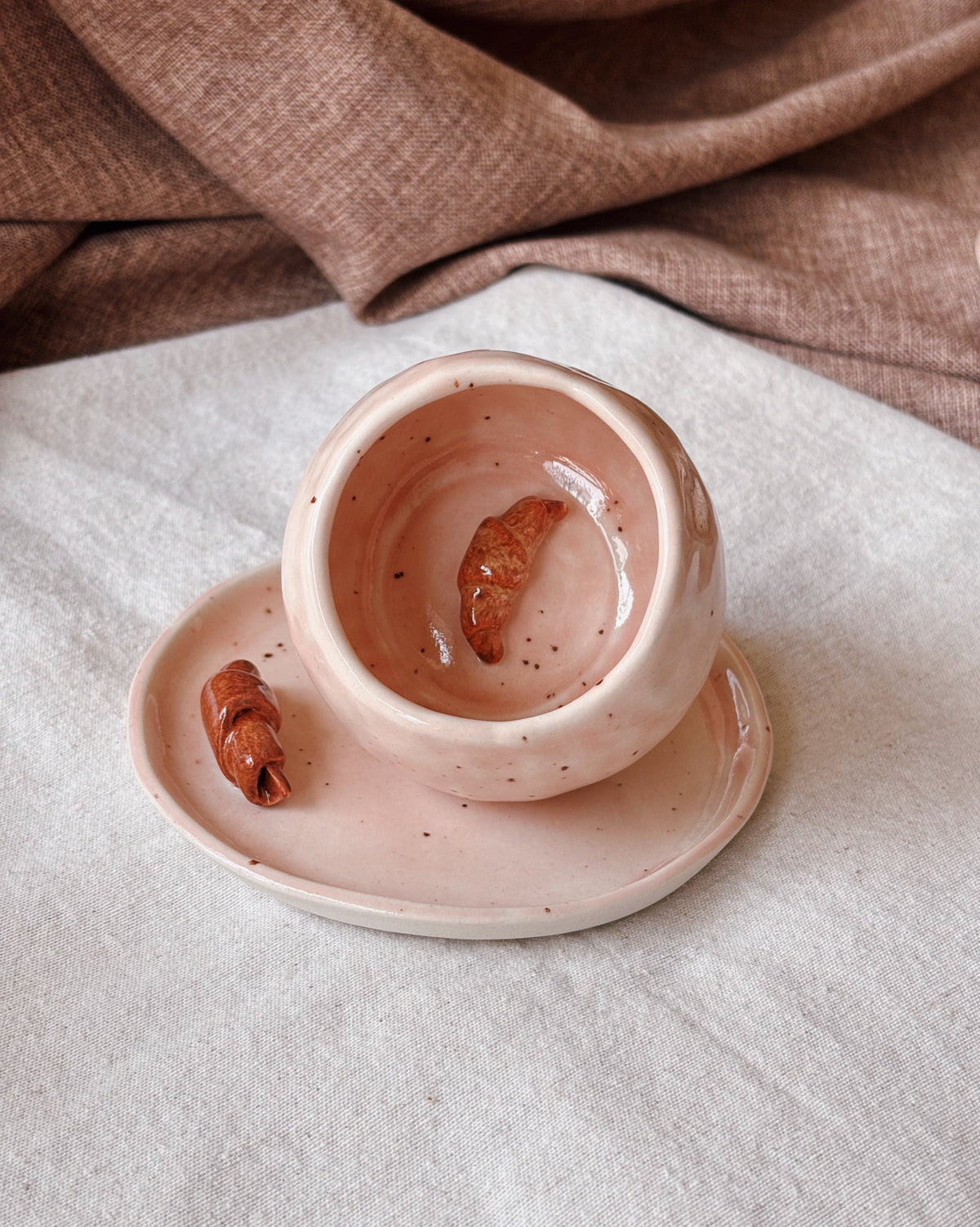 A small pink ceramic plate with a matching mug, both speckled with dark flecks, styled on a light fabric surface with a draped brown textile in the background.