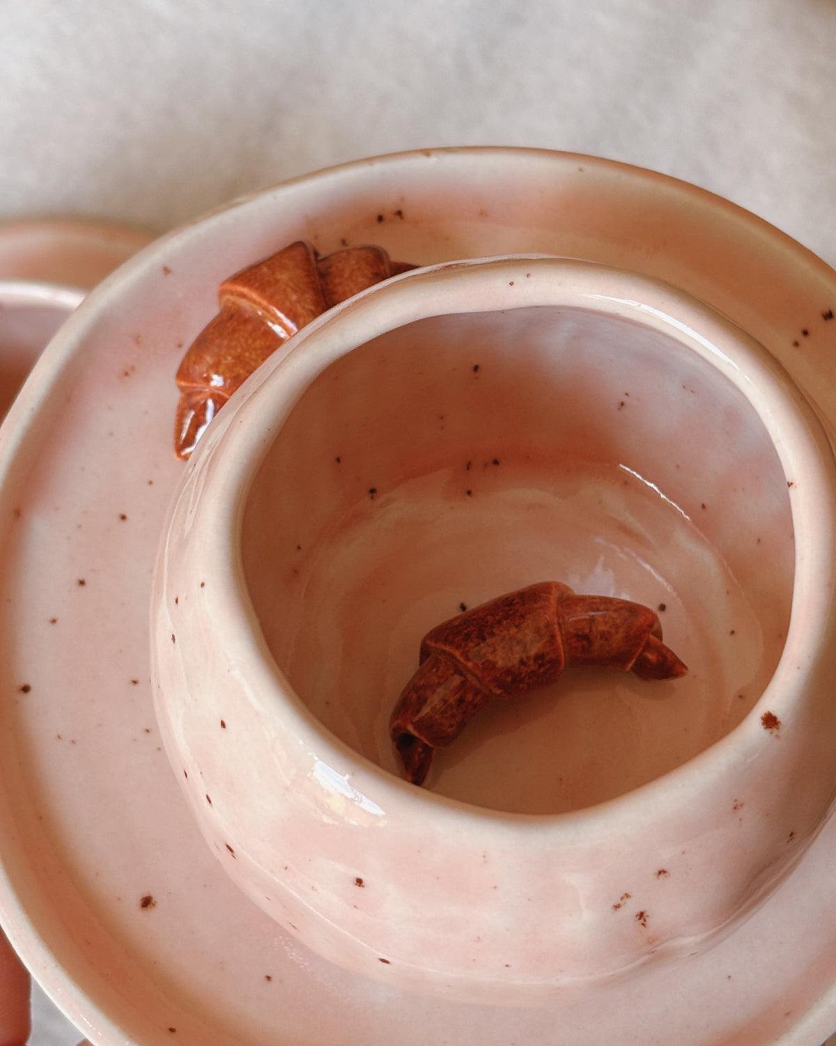 A close-up of a light pink speckled ceramic cup or bowl with a small brown croissant inside, set against a soft neutral background.