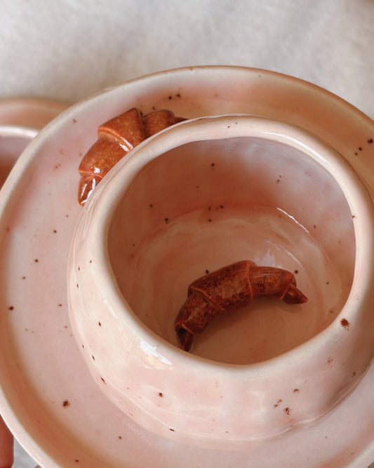 A close-up of a light pink speckled ceramic cup or bowl with a small brown croissant inside, set against a soft neutral background.