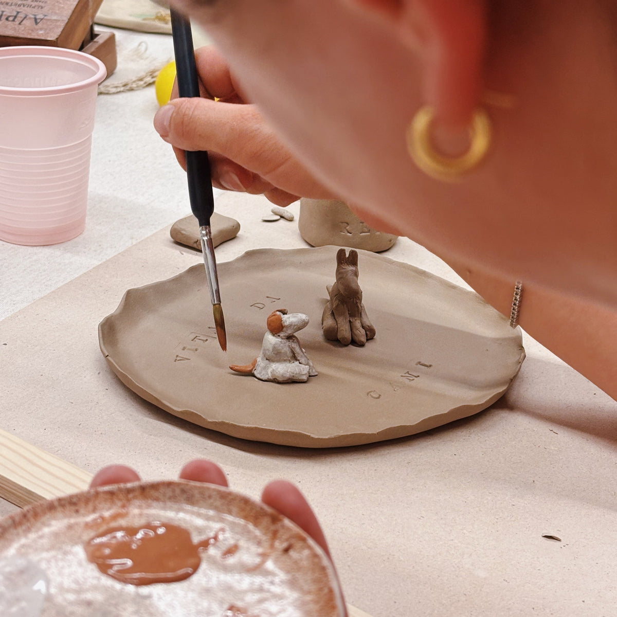 A close-up lifestyle shot of a person painting miniature clay figurines on a small clay plate. The person is holding a paintbrush and a small dish of brown paint in a pottery studio setting.