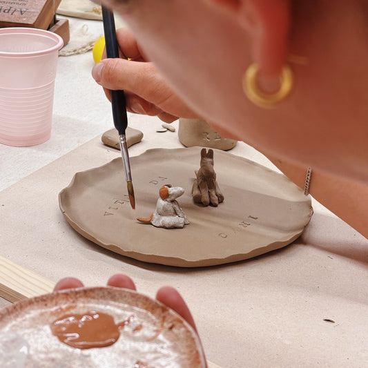 A close-up lifestyle shot of a person painting miniature clay figurines on a small clay plate. The person is holding a paintbrush and a small dish of brown paint in a pottery studio setting.