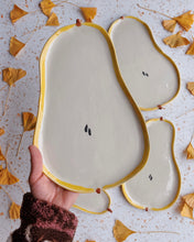 Load image into Gallery viewer, Hand holding a pear-shaped ceramic plate with gold edges on a textured surface with autumn leaves.
