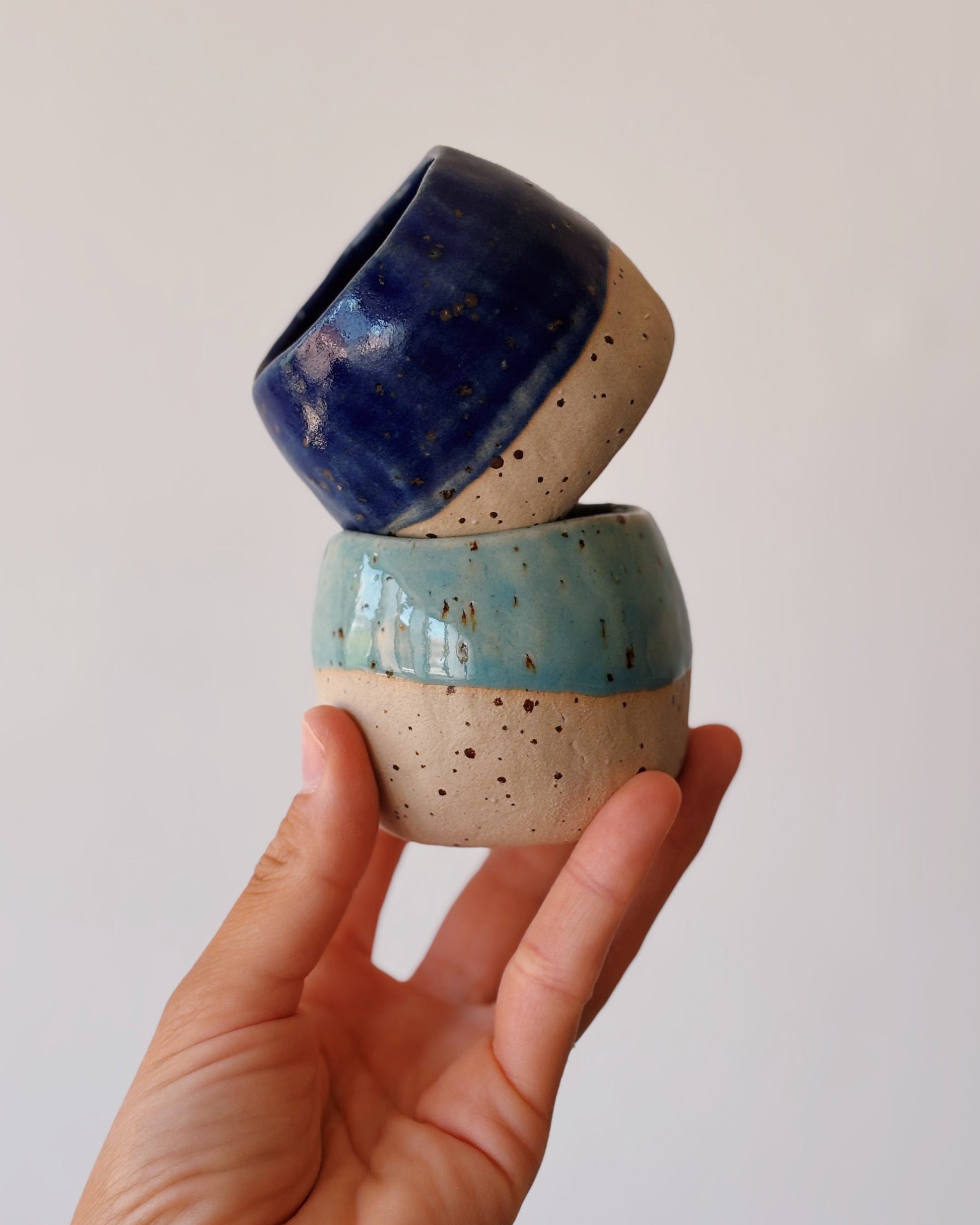 A hand holding two small handmade ceramic cups stacked on top of each other against a plain light background. The cups have a speckled cream base with a light blue glazed section on the lower cup and a dark blue glazed section on the upper cup.