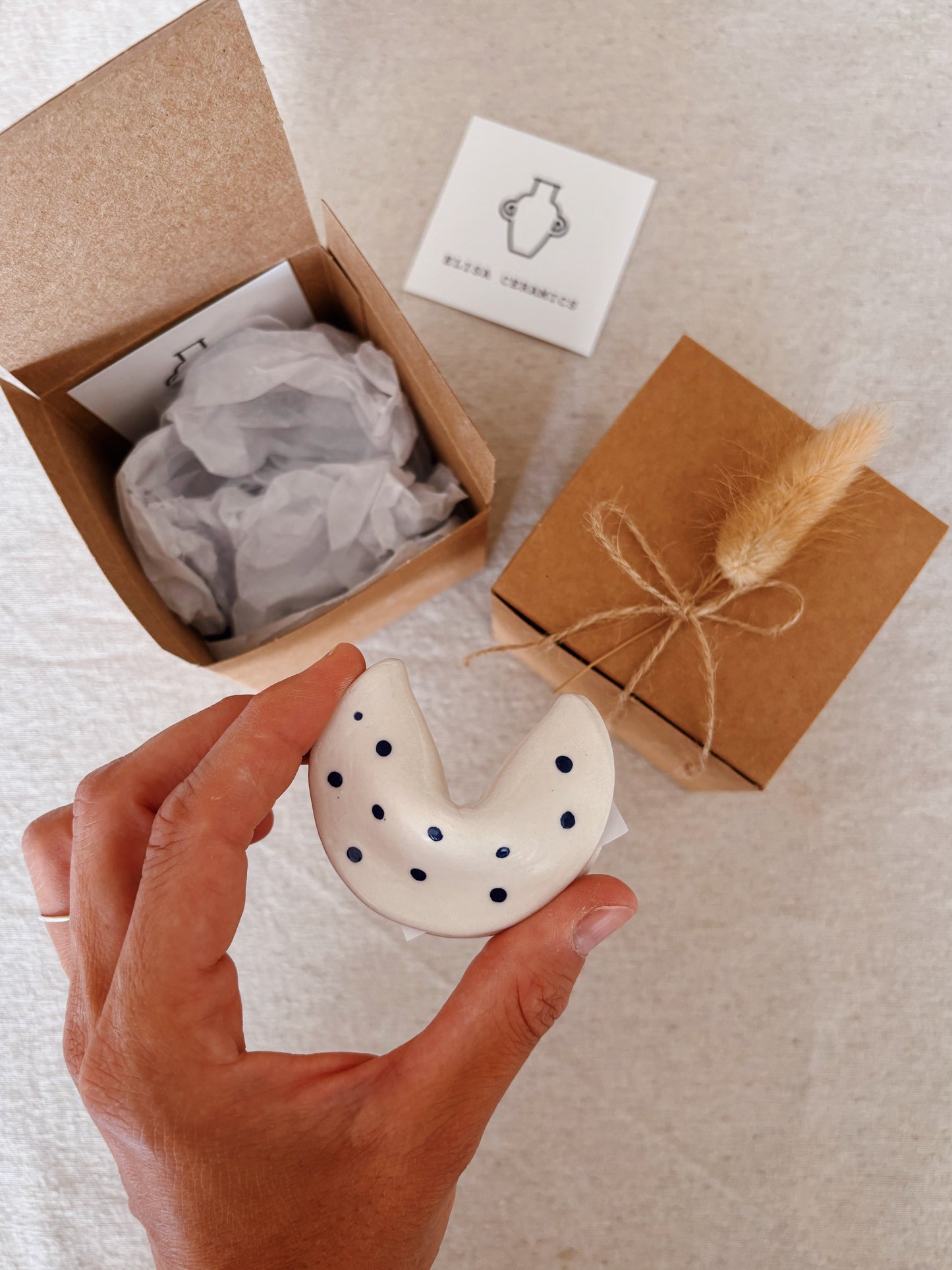 A hand holds a small handmade ceramic fortune cookie with dark blue polka dots, shown next to kraft paper gift boxes and a small branded card on a light fabric surface.