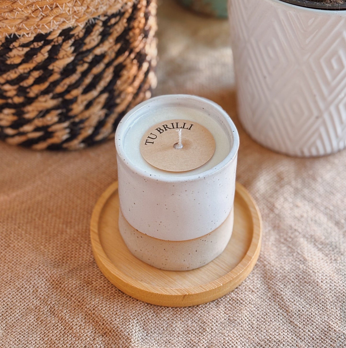 White candle with wooden lid on a coaster, surrounded by potted plants on a textured surface