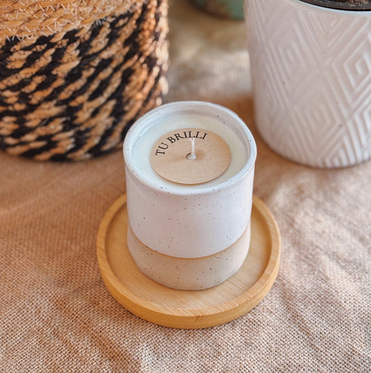 White candle with wooden lid on a coaster, surrounded by potted plants on a textured surface
