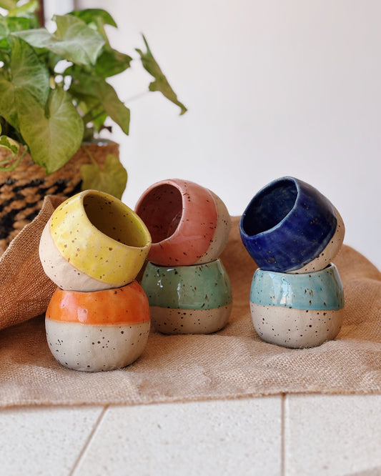 Six handmade ceramic coffee cups stacked in pairs of two, displayed on a beige table runner with a potted green plant in the background.