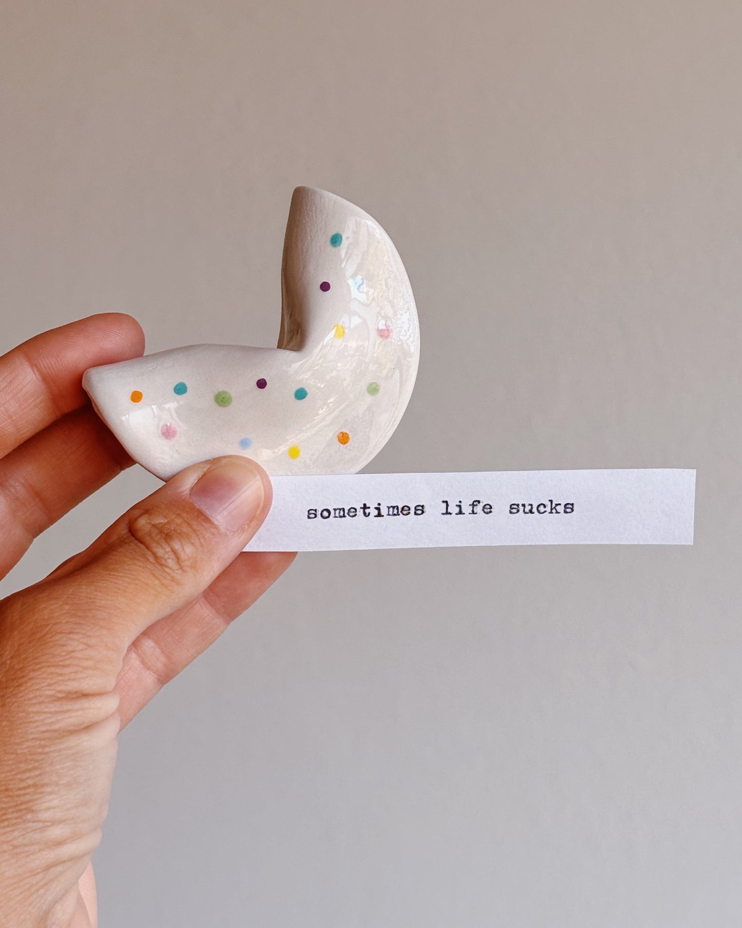A hand holds a small ceramic fortune cookie decorated with colorful polka dots next to a white paper strip printed with the text 'sometimes life sucks' against a plain light gray background.