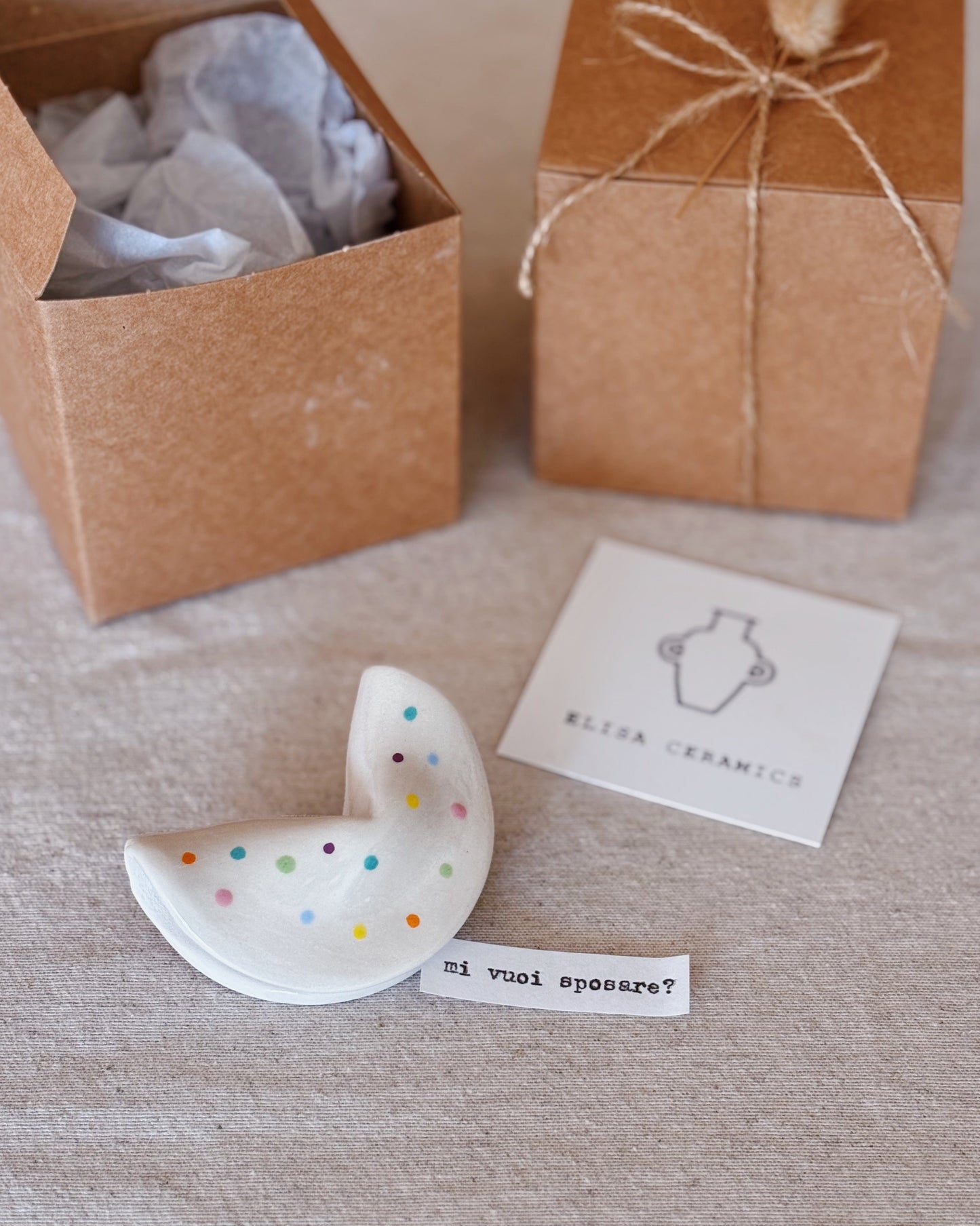 A white ceramic fortune cookie with colorful polka dots, displayed next to a small white card reading 'mi vuoi sposare?'. In the background are two brown kraft gift boxes on a beige linen surface.