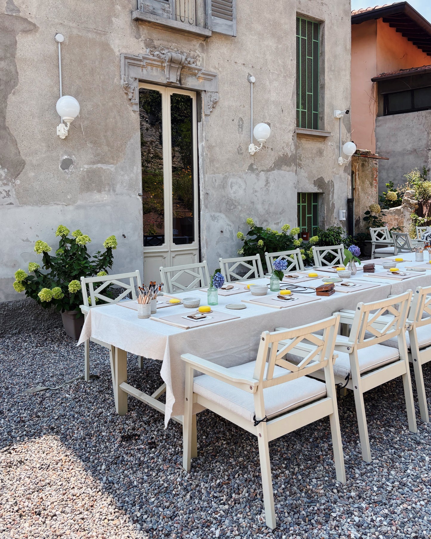An outdoor ceramic workshop setup on a gravel courtyard in front of a rustic stone building, featuring a long table with a white tablecloth and multiple cream-colored wooden chairs with white cushions.
