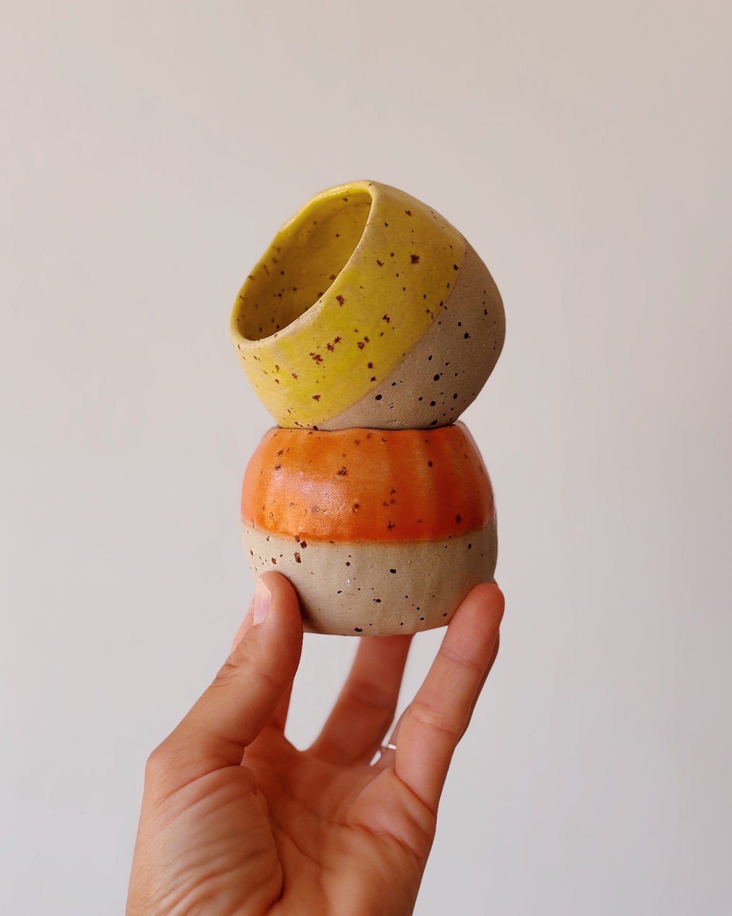 A hand holding a small ceramic stack of two speckled bowls, one yellow and one orange and cream, against a plain light background.
