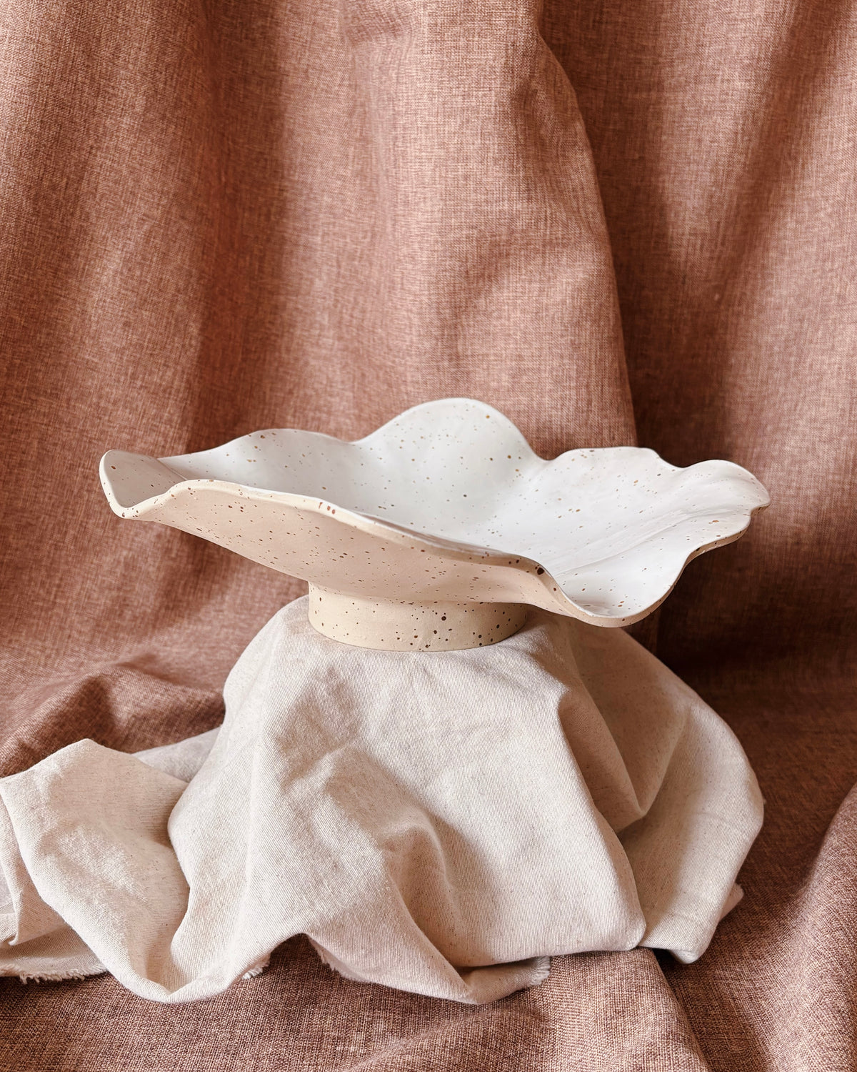 A handmade ceramic vase with a wavy rim, speckled with small dark flecks, resting on draped beige linen fabric against a matching brown linen backdrop.