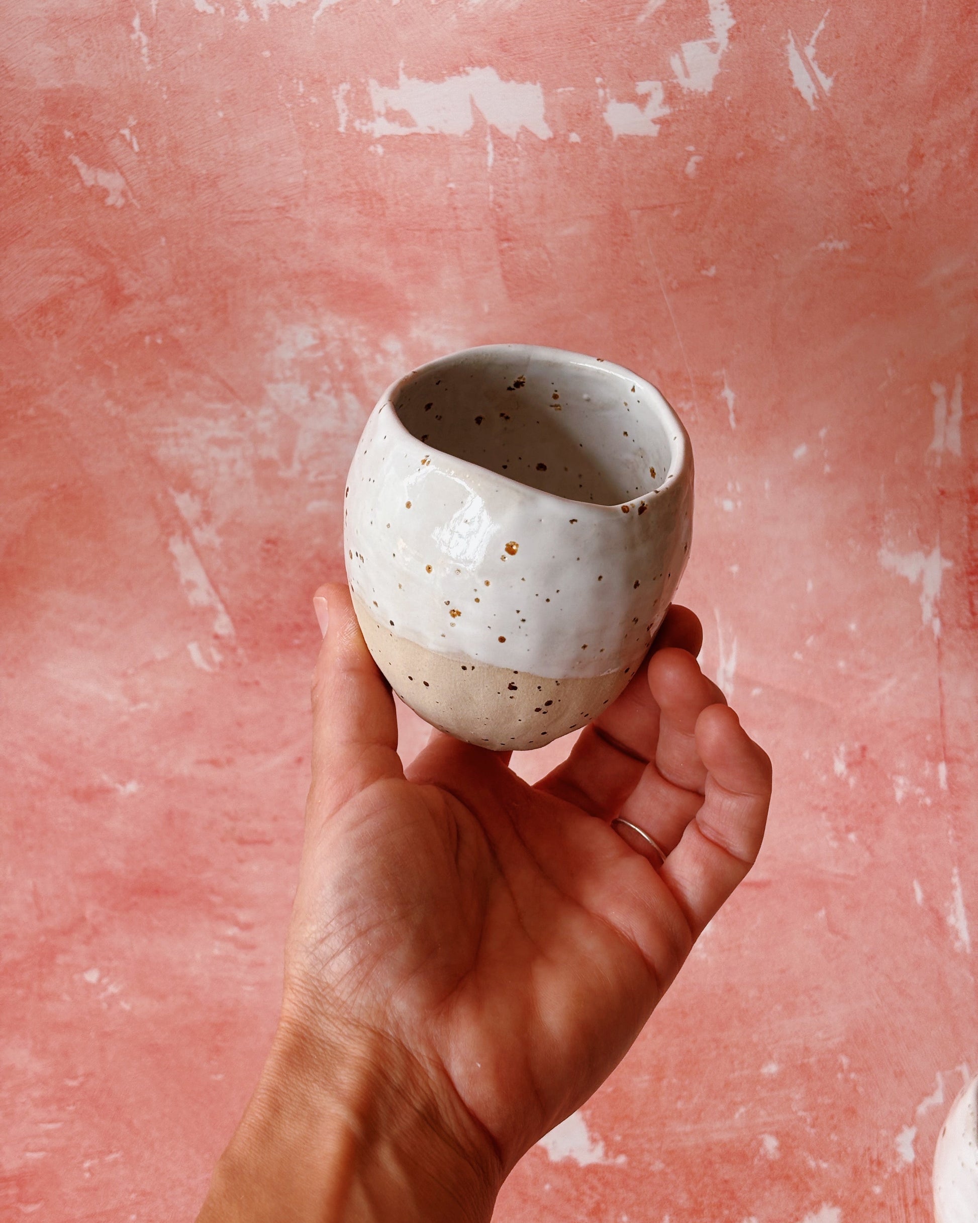 Hand holding a small speckled ceramic cup against a pink textured background
