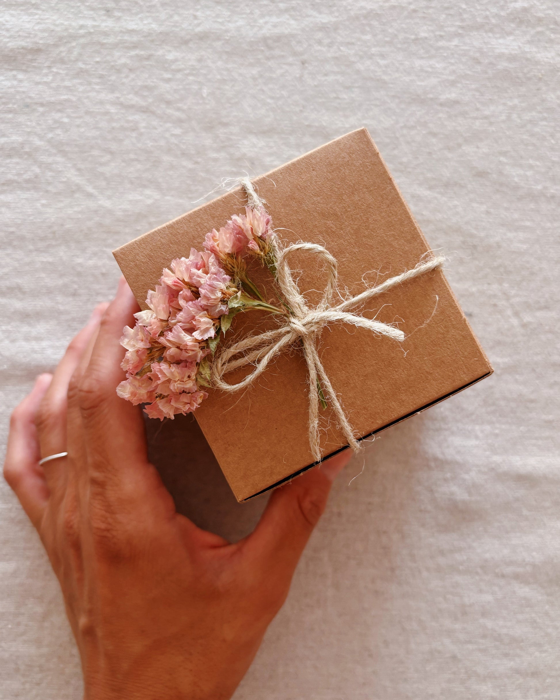 A hand holding a small square kraft paper gift box tied with natural twine and decorated with dried pink flowers, set against a textured off-white fabric background.