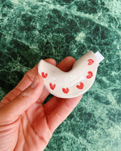 Load image into Gallery viewer, Hand holding a small ceramic fortune cookie with red hearts against a green marble background
