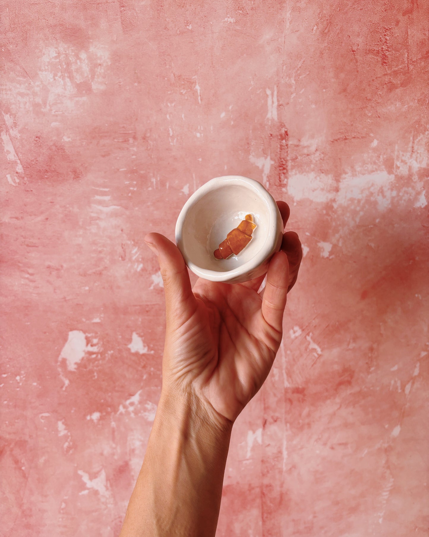 Hand holding a handmade stoneware croissant espresso mug against a pink background.