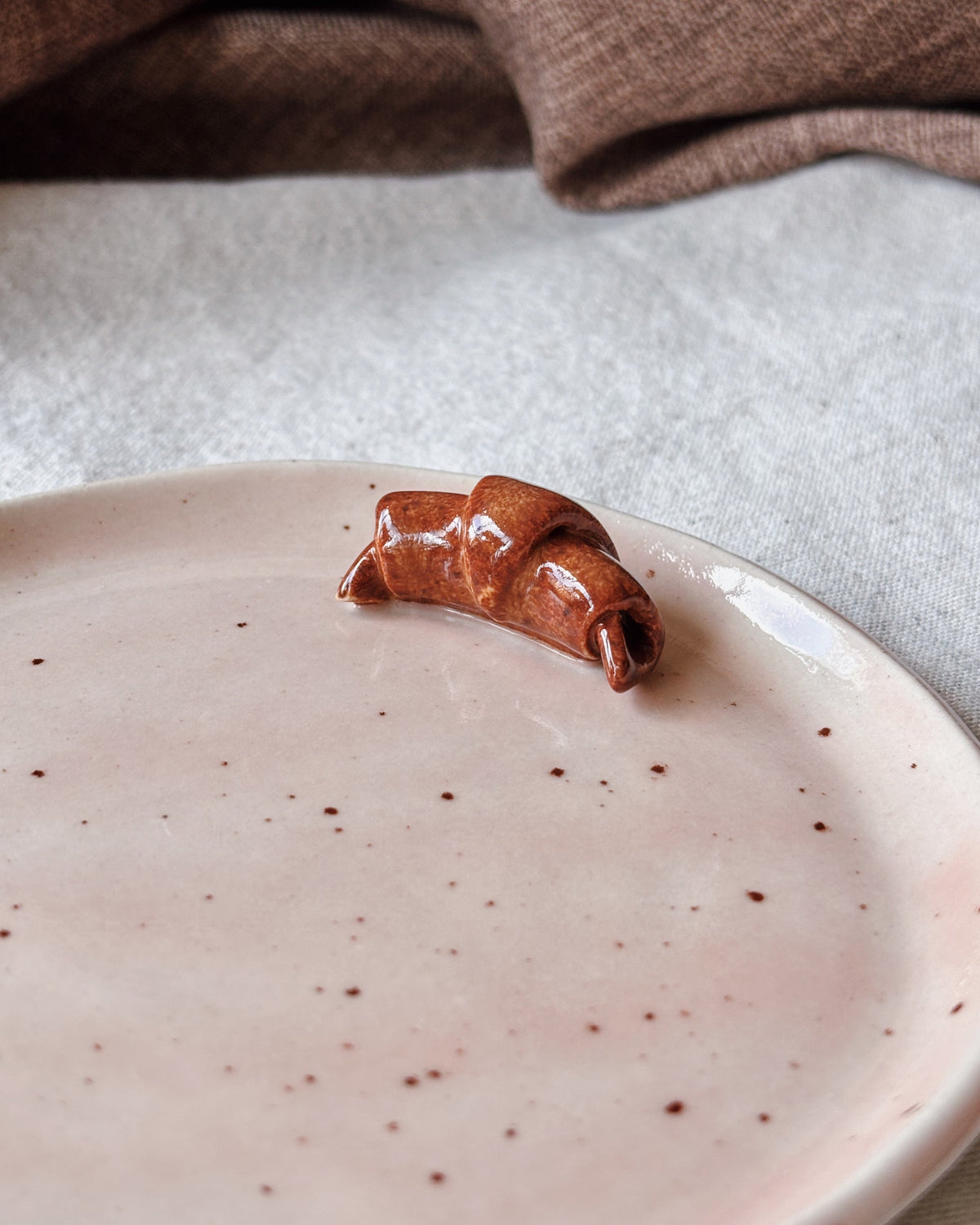 A small glossy brown ceramic croissant-shaped object sits on a pale pink speckled plate, with a neutral fabric surface and a brown textile draped in the background.