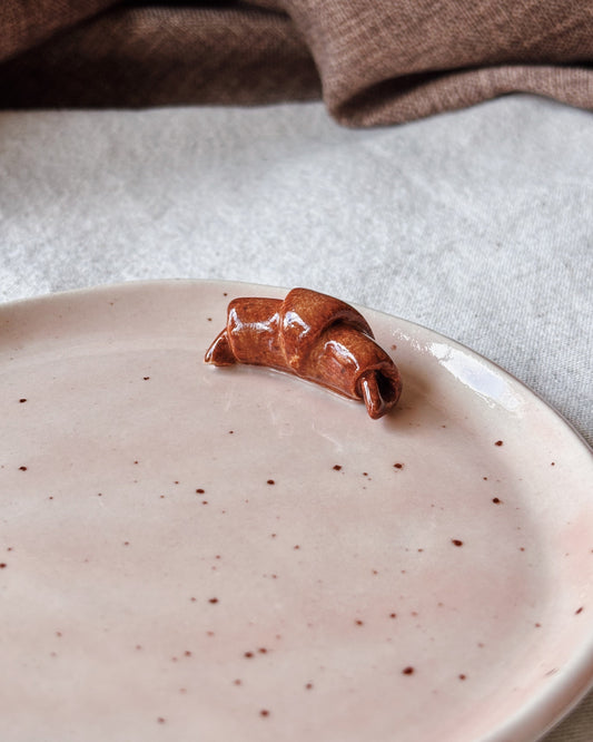 A small glossy brown ceramic croissant-shaped object sits on a pale pink speckled plate, with a neutral fabric surface and a brown textile draped in the background.