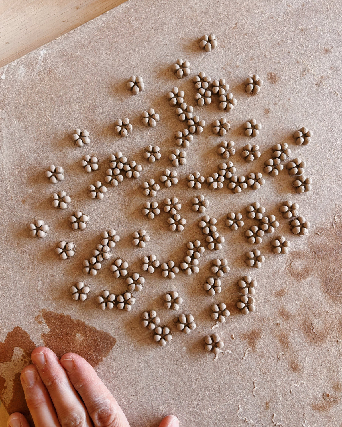 Small clay flower appliqués scattered on brown parchment paper, with a hand visible at the bottom left and a small knife at the top on a wooden surface.
