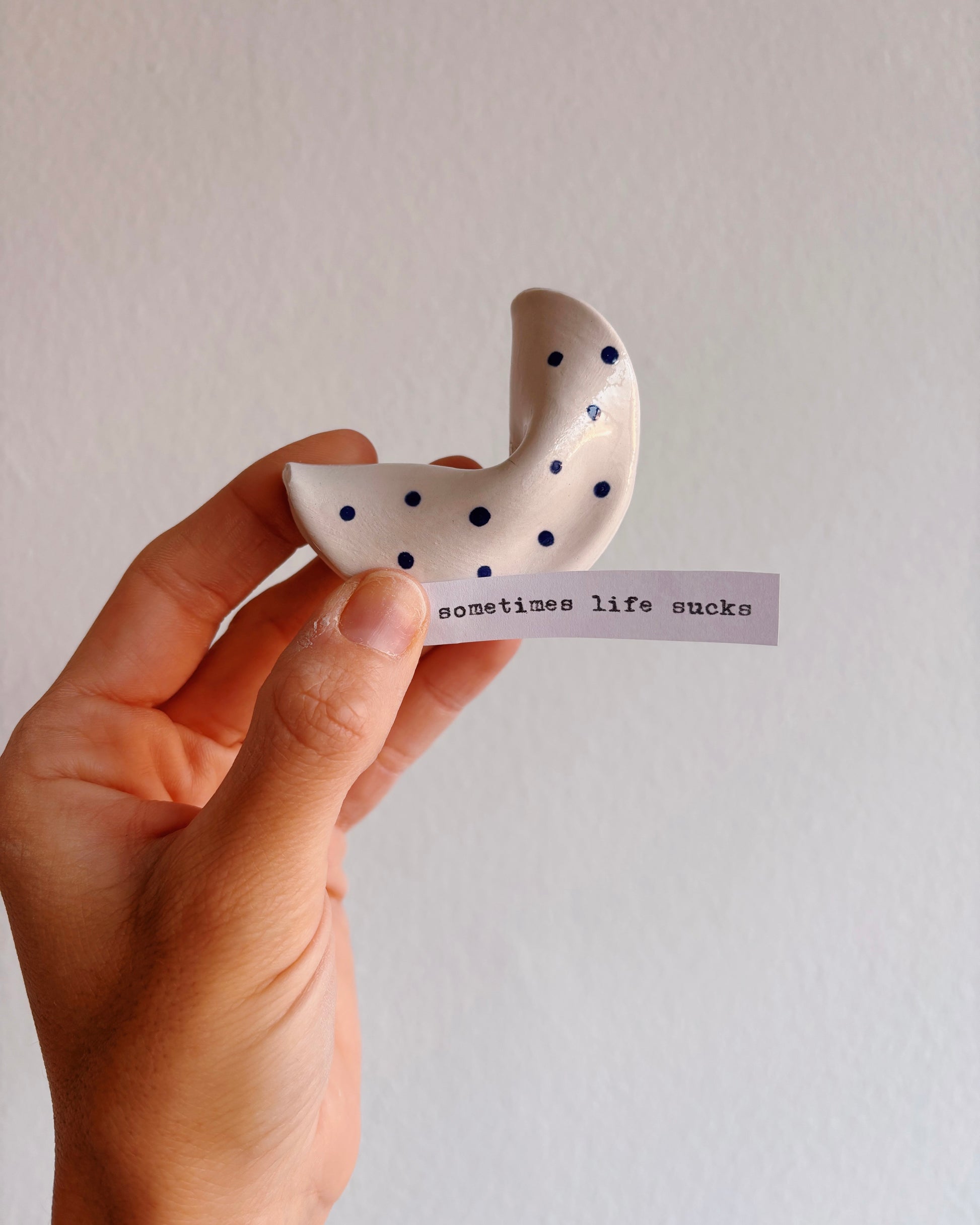 Hand holding a white ceramic crescent moon with black dots and a small slip of paper 'sometimes life sucks' on a plain background.