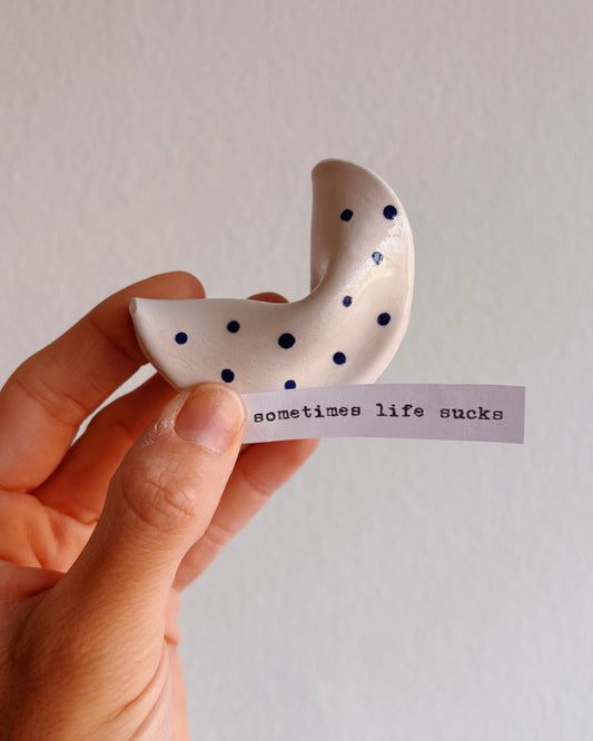 Hand holding a white ceramic crescent moon with black dots and a small slip of paper 'sometimes life sucks' on a plain background.