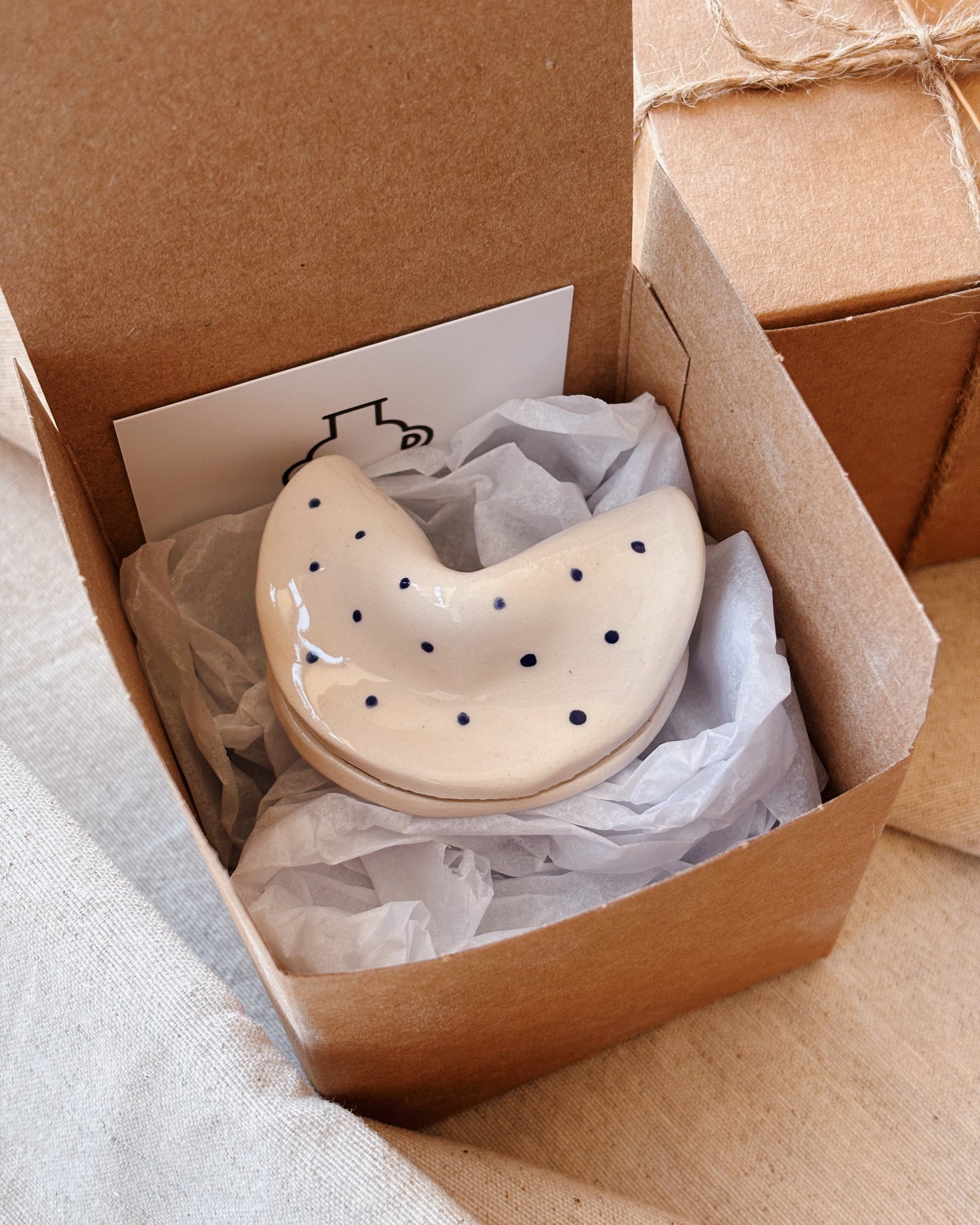 A cream-colored ceramic fortune cookie with small black polka dots, nestled in white tissue paper inside an open brown kraft gift box.