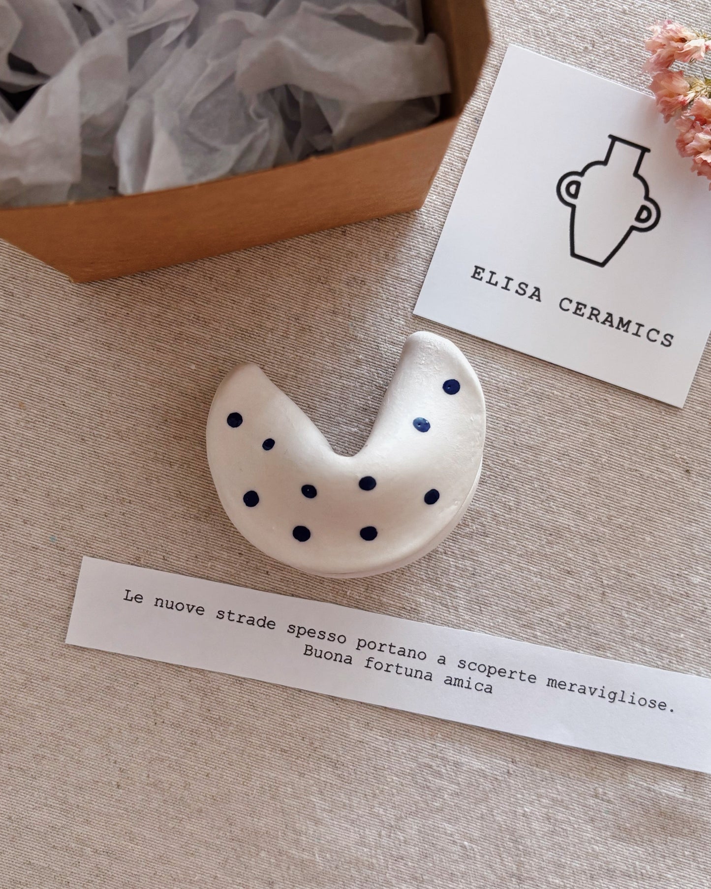 A ceramic fortune cookie  with black polka dots on a white base, displayed next to a brand card for Elisa Ceramics and a handwritten note on a beige fabric surface next to an open gift box.