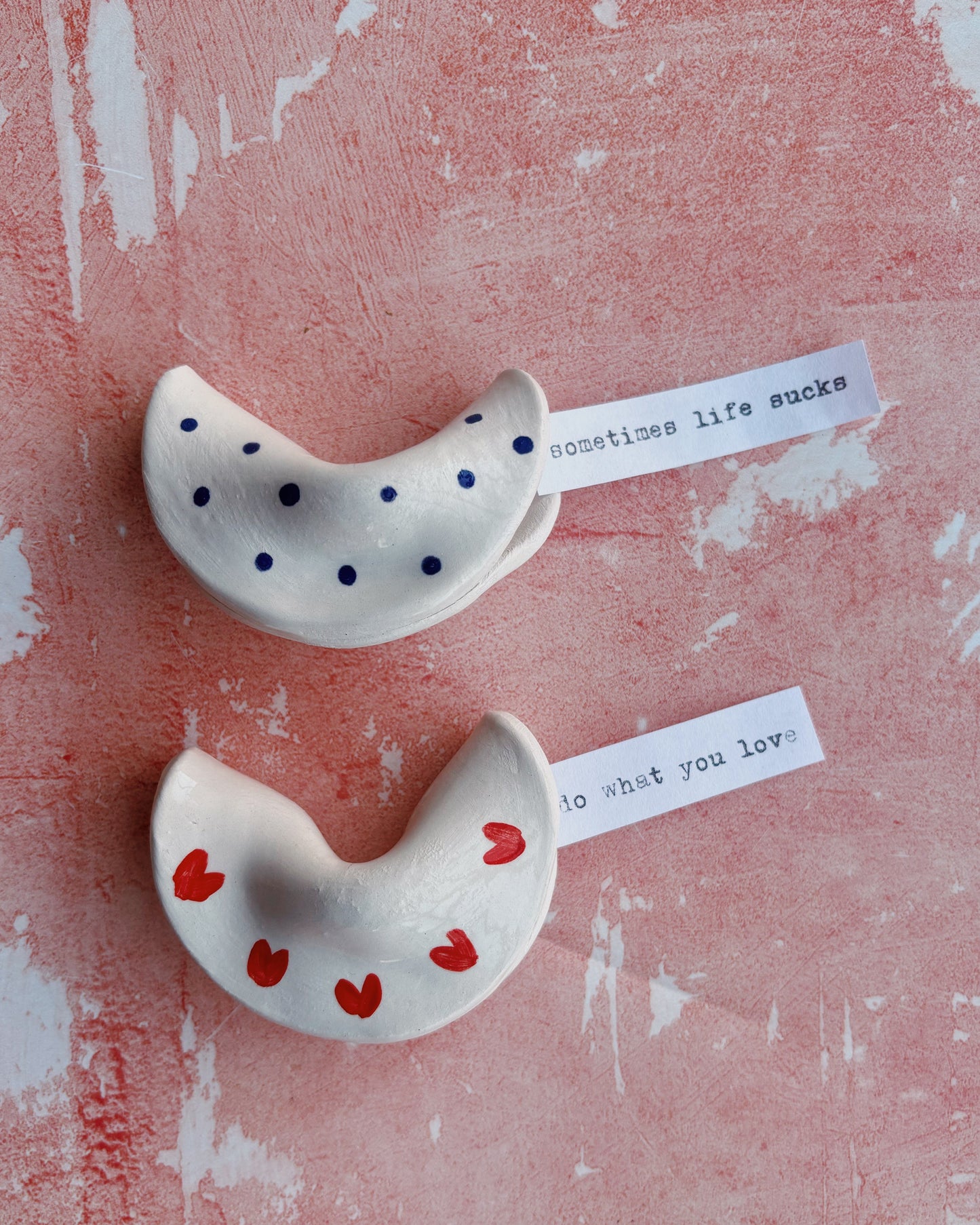 Two ceramic fortune cookies with polka dots and hearts on a textured pink surface.