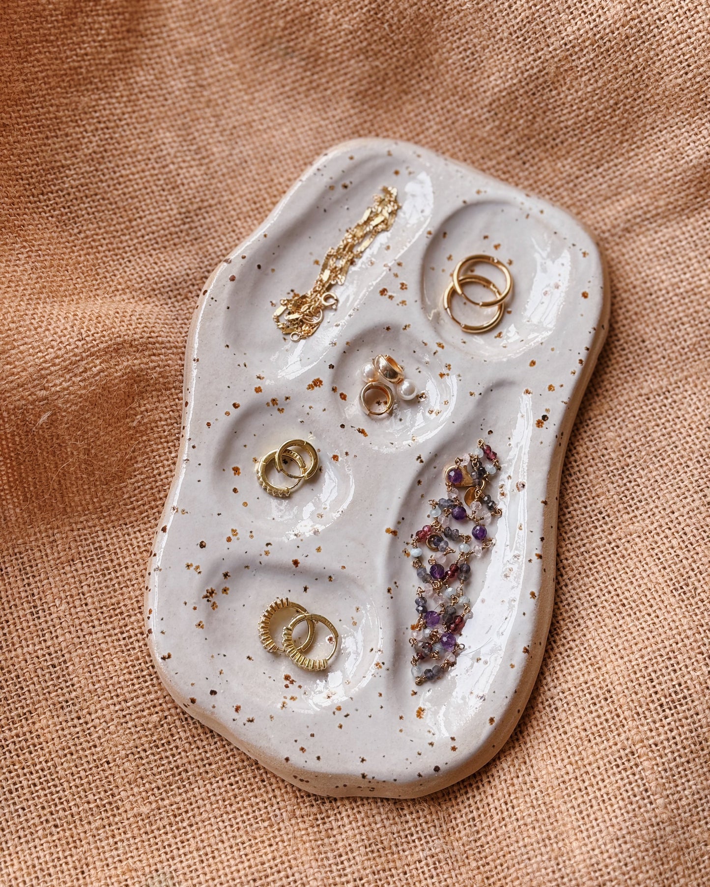 Speckled ceramic jewelry dish with gold and silver jewelry on a beige fabric background