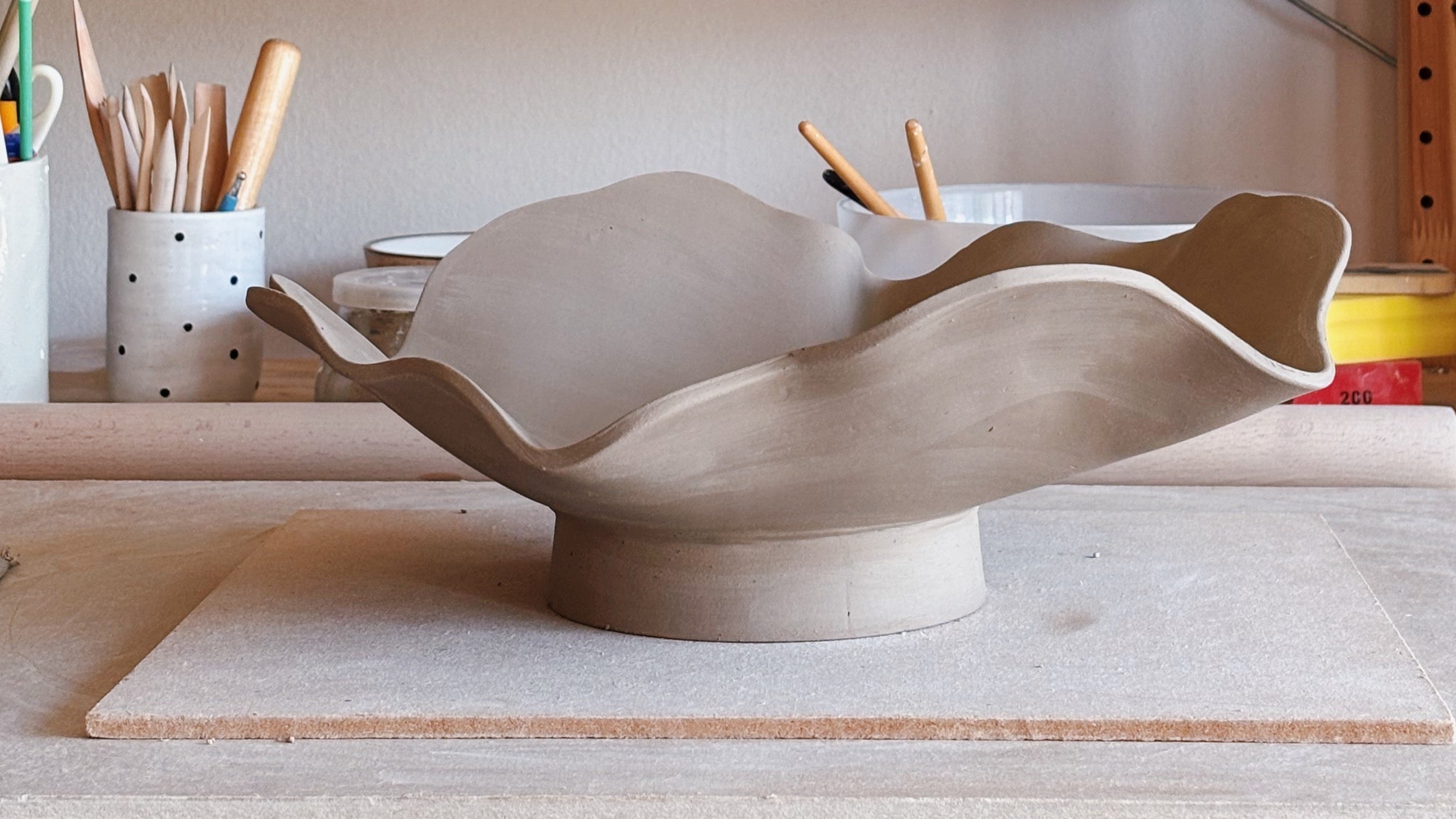 A handmade ceramic flower-shaped bowl in a matte light beige clay finish, displayed on a wooden workbench inside a pottery studio with shelves of pottery tools and plants in the background.