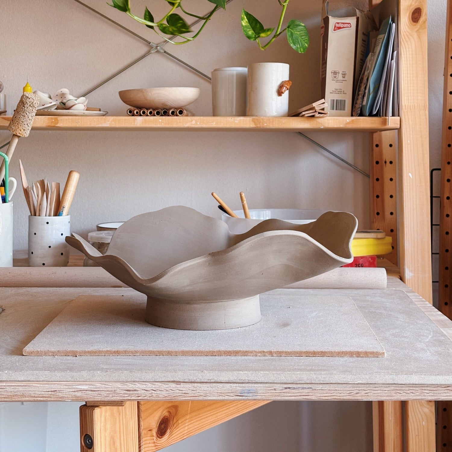 A handmade ceramic flower-shaped bowl in a matte light beige clay finish, displayed on a wooden workbench inside a pottery studio with shelves of pottery tools and plants in the background.