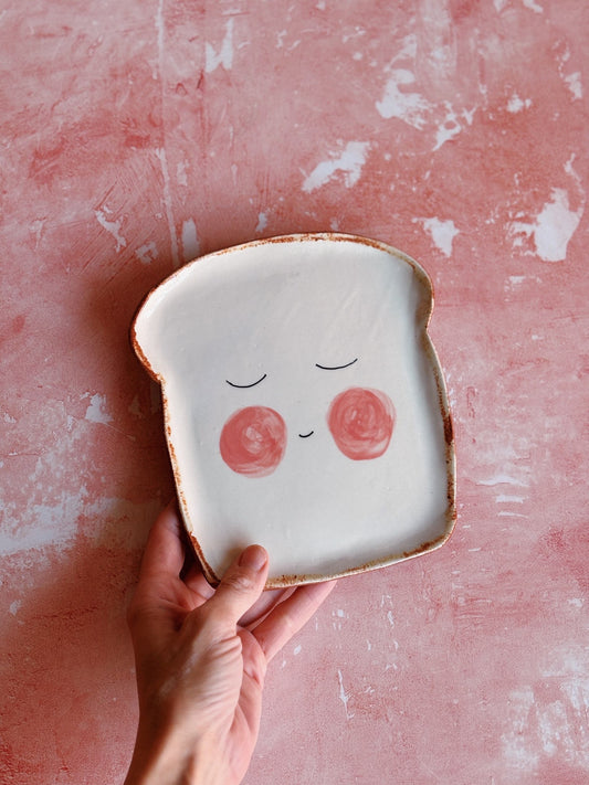 Hand holding a ceramic plate shaped like a slice of bread with a face on a pink textured background