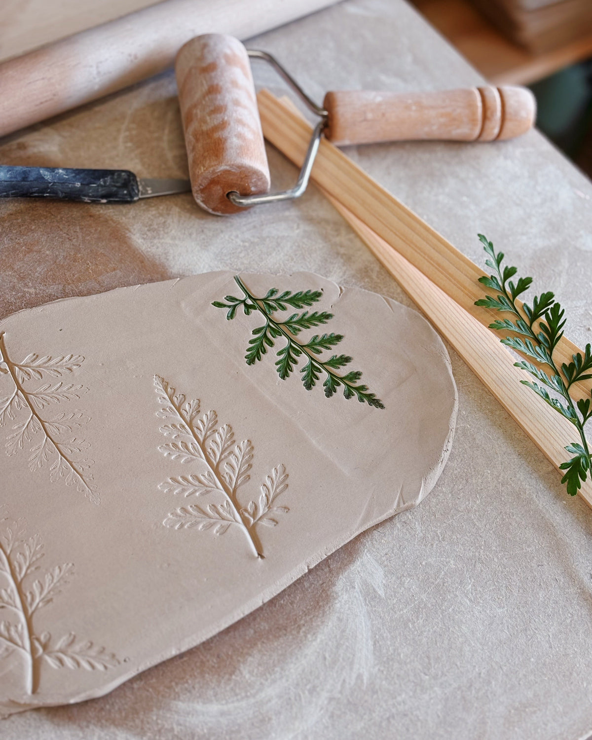 A close-up of a beige clay slab stamped with leaf and fern patterns, with green pressed foliage placed on top. A wooden rolling pin, a small roller tool, and wooden tools rest on a light surface in a styled craft setting.