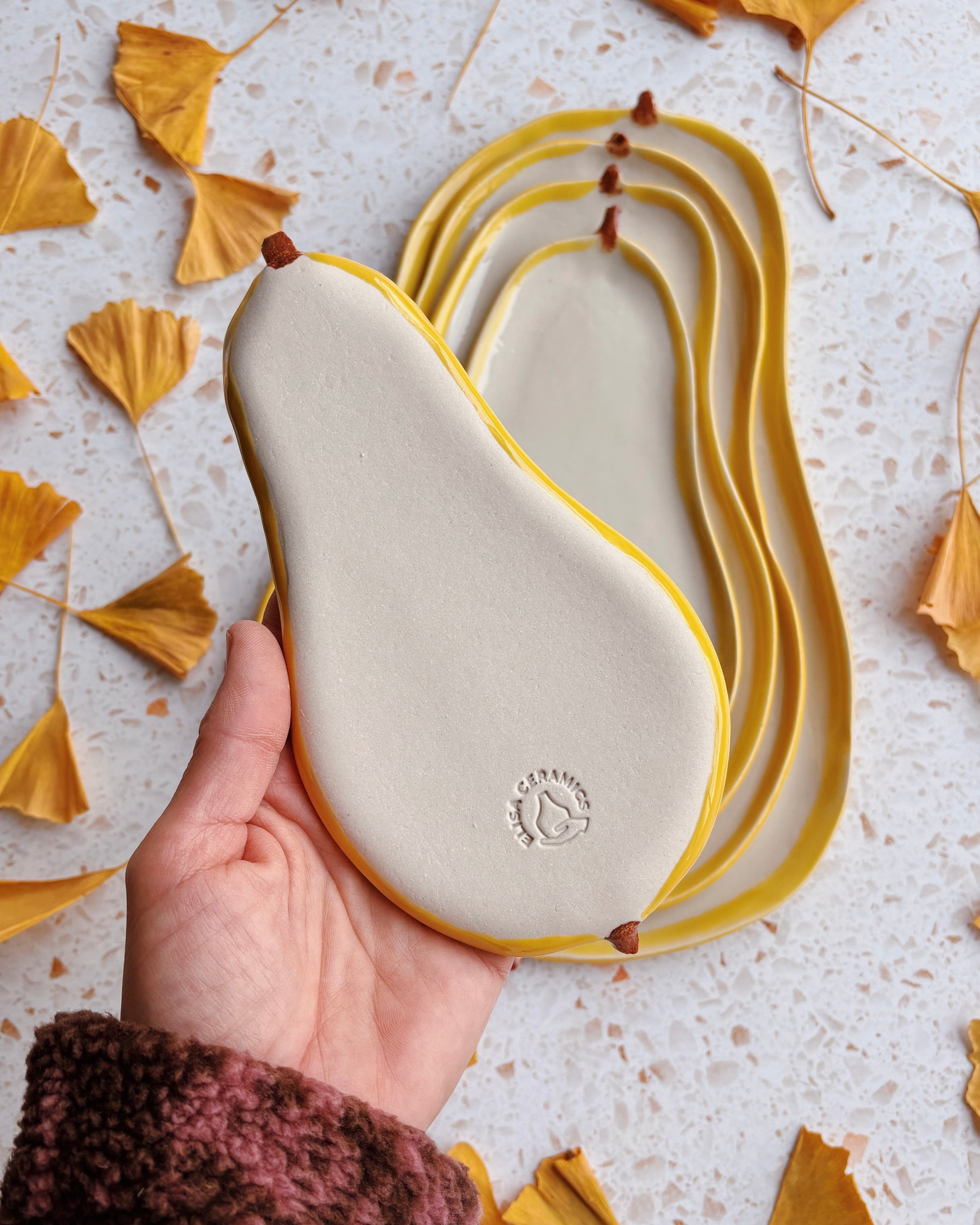 Hand holding a ceramic pear-shaped plate with a yellow rim on a light background with autumn leaves.