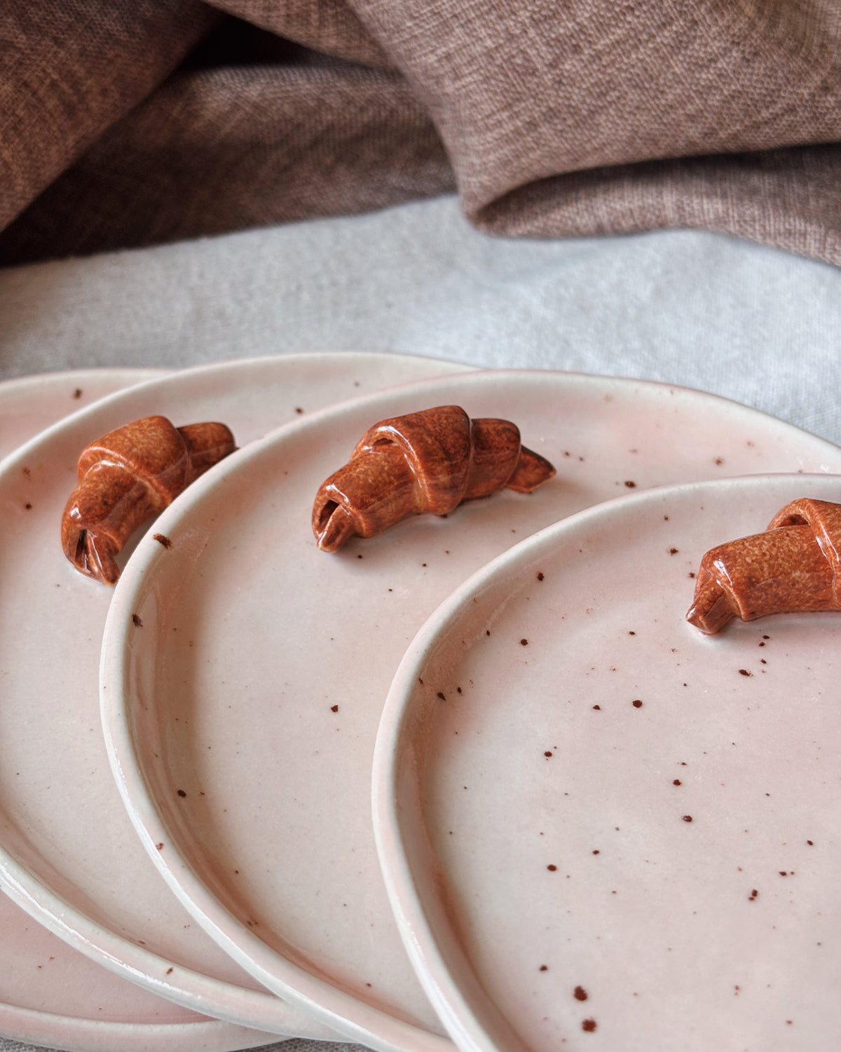 Three pale pink speckled ceramic plates arranged in an overlapping fan, each holding a small croissant-shaped pastry, with a soft brown fabric draped in the background.