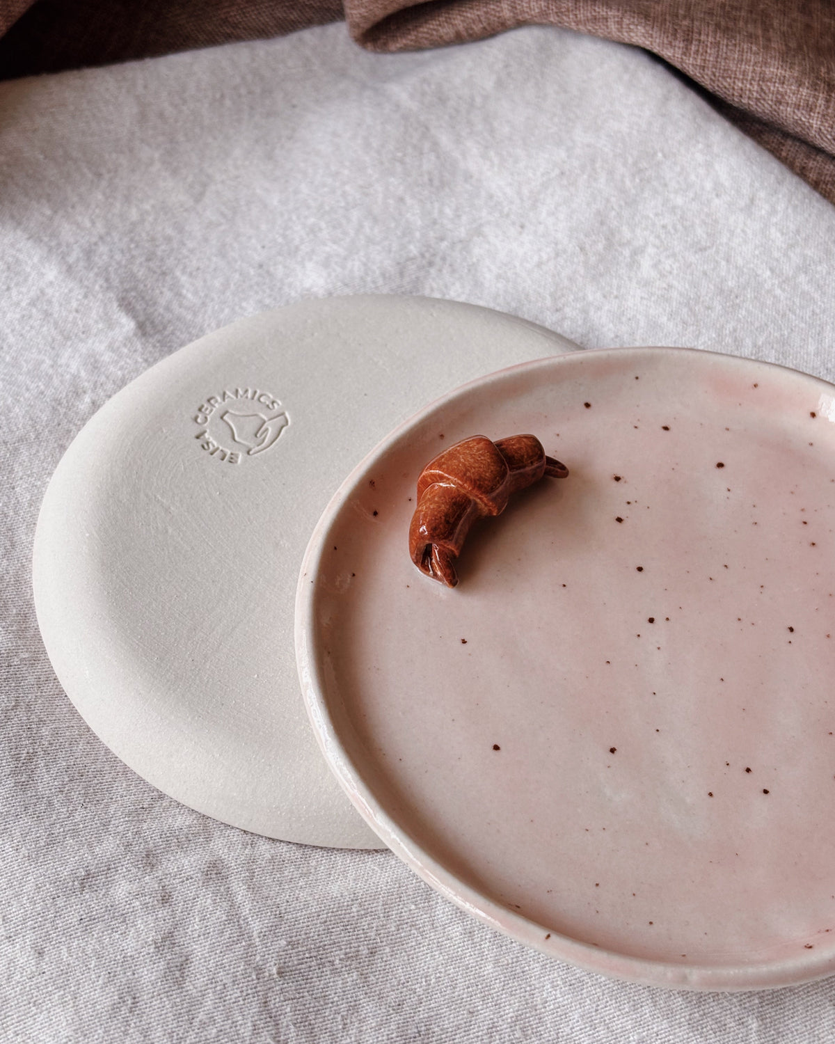 Two handmade ceramic plates in a neutral studio setting. One plate is shown from the back, displaying a matte off-white finish and a debossed circular logo, while the other is shown from the front in a soft blush pink color with tiny dark speckles, styled with a small brown pastry on top.
