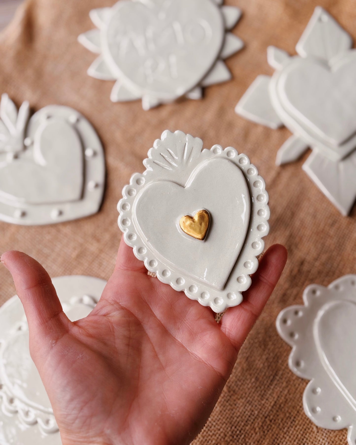 Hand holding a white ceramic heart-shaped object with a gold heart inset on a brown surface with more ceramic pieces.