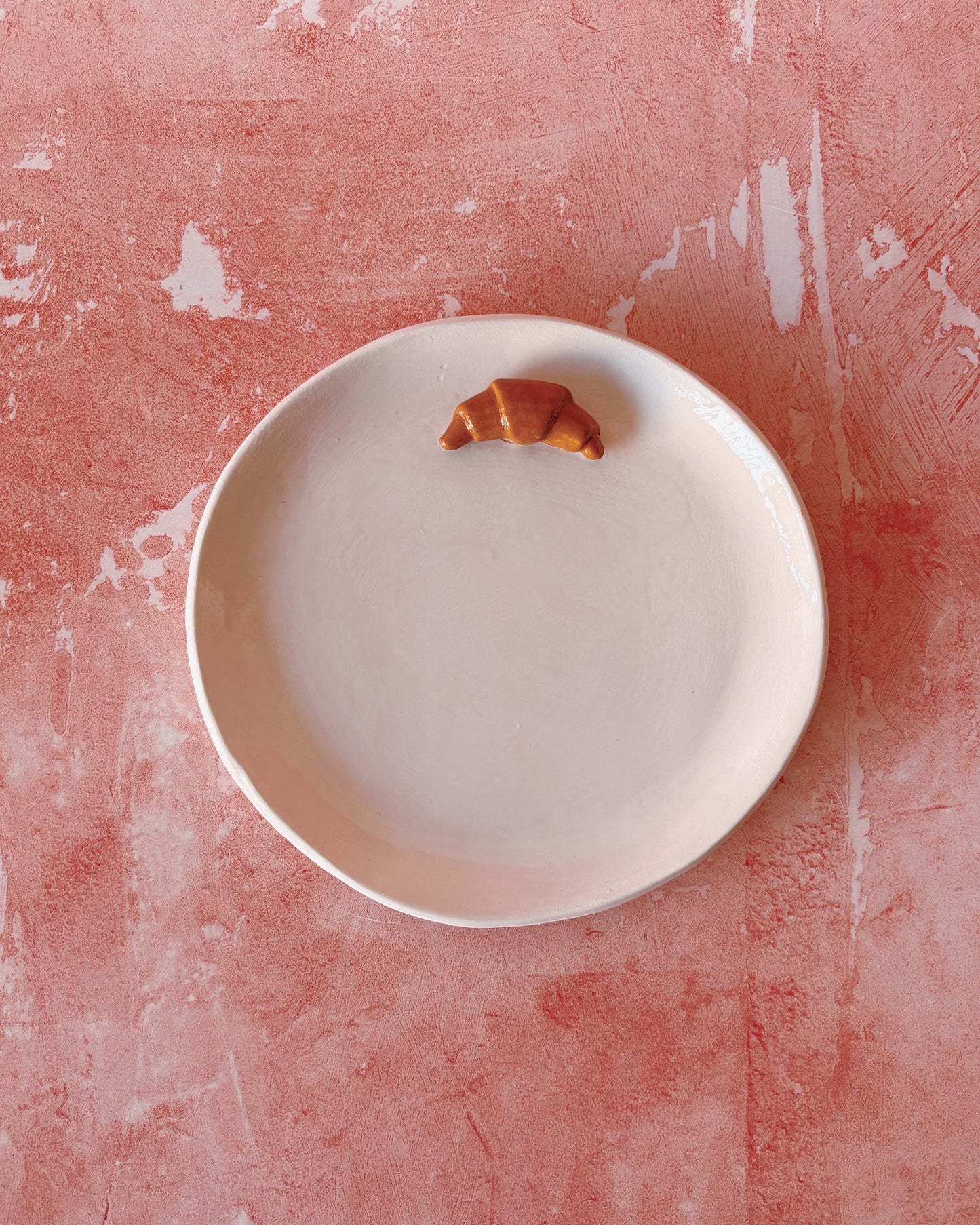 Croissant Small Ceramic Plate with a croissant on a textured pink background, handmade stoneware for breakfast use.