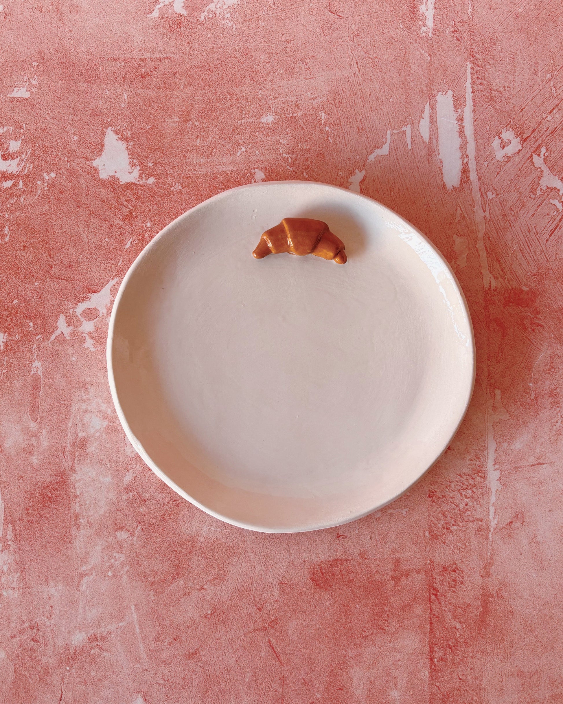 Croissant Small Ceramic Plate with a croissant on a textured pink background, handmade stoneware for breakfast use.