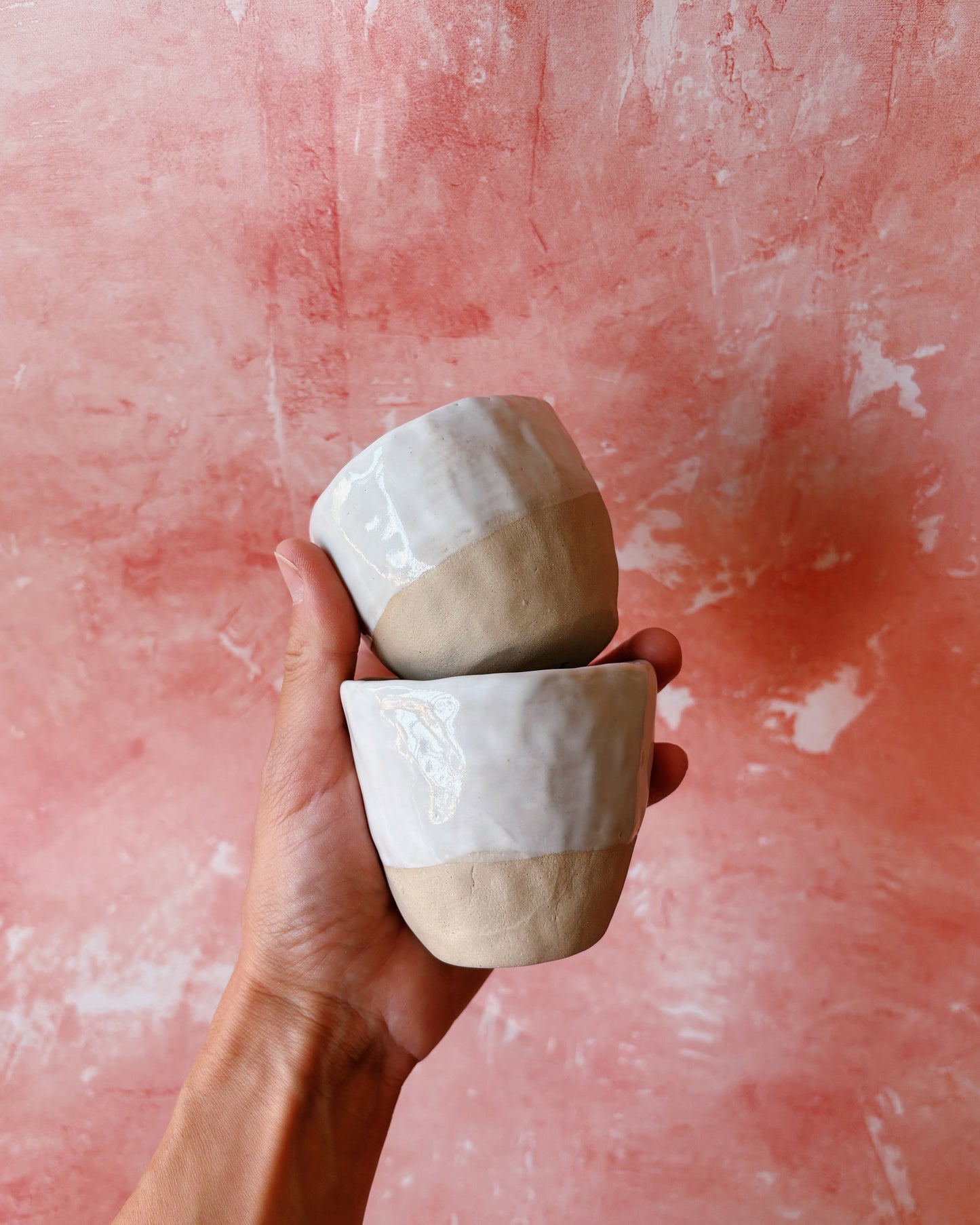 Two ceramic cups with a two-tone design held against a pink textured background