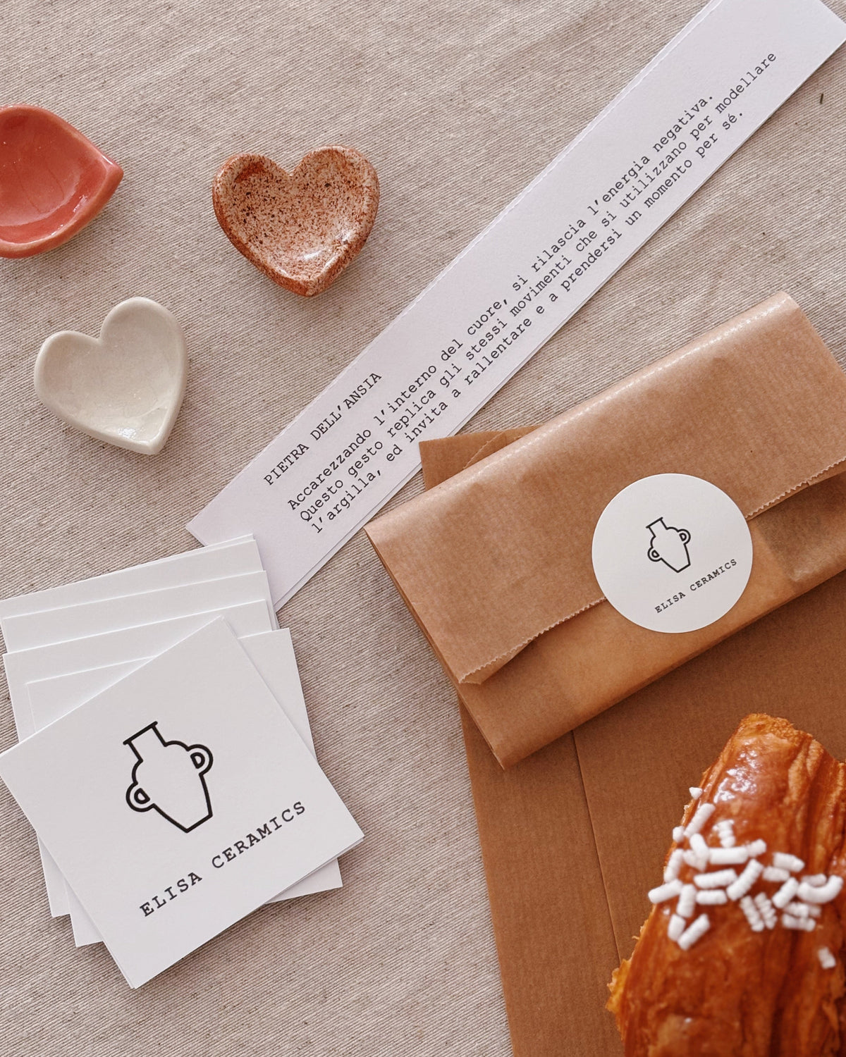 A styled flat-lay of a ceramic product set by Elisa Ceramics, including a brown kraft paper envelope with a round white logo sticker, a stack of white square cards with the same logo, a long white printed card, and a croissant on a brown paper sheet, all arranged on a beige fabric background.