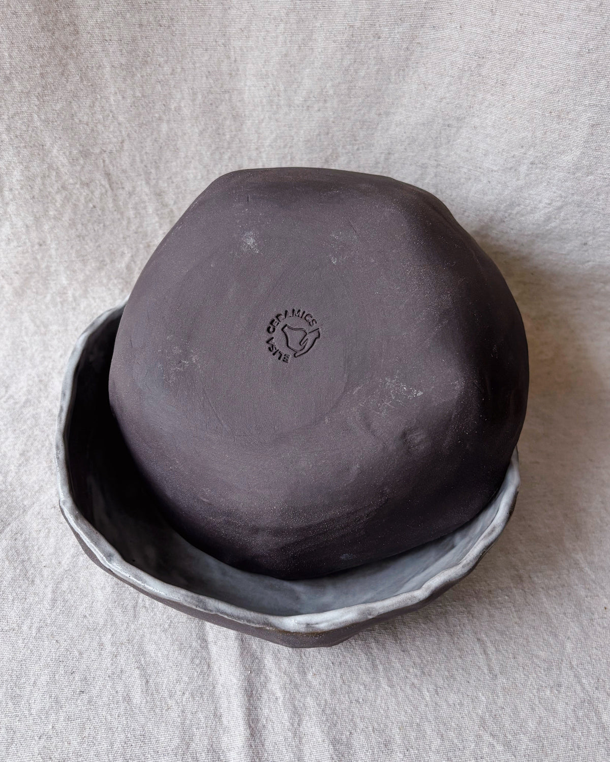 A handmade dark gray ceramic bowl with a textured, matte finish, shown upside down in a shallow matching bowl on a light beige fabric background.