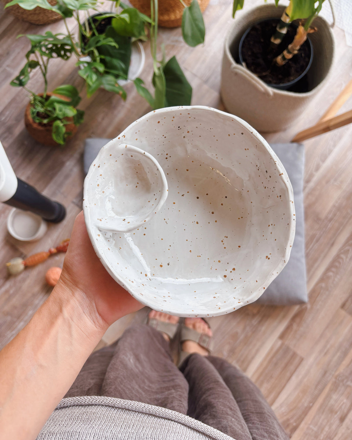 A handmade ceramic bowl with a small integrated cup, held by a hand in a cozy indoor setting with wooden floors and potted plants in the background.