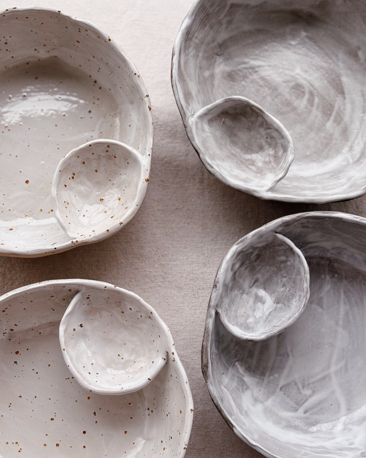 Four handmade ceramic bowls with integrated spoons, arranged on a neutral textured surface. Three bowls are off-white with subtle brown speckles, and one is a matte gray.